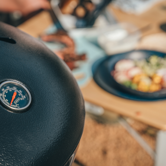 Cadac Carri Chef 40 - BBQ & Chef Pan Combo lifestyle image close up of the thermometer in the lid with out of focus food on the table in the background. 