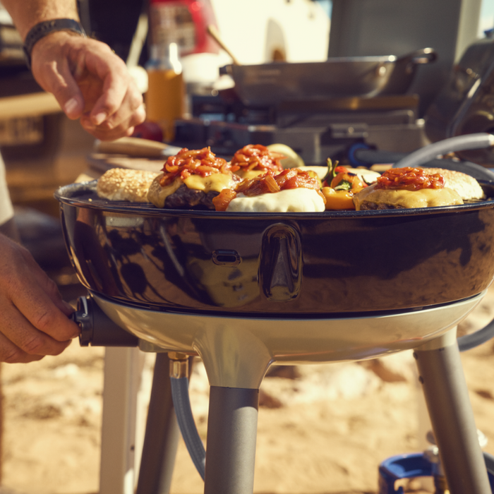 Cadac Carri Chef 40 - BBQ & Chef Pan Combo lifestyle image of a hand turning the gas as burgers and veg cook on top of the grill a cadac stove can be seen in the background with pan on stove. 
