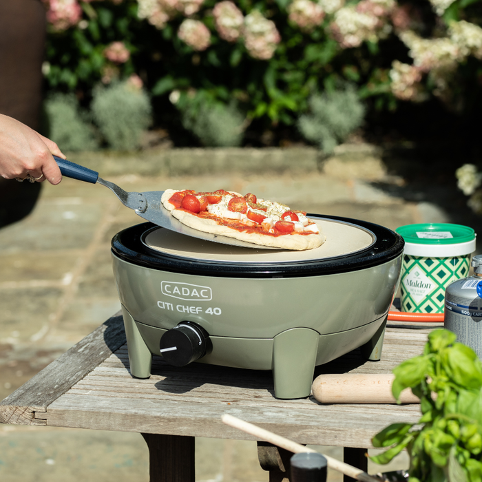 Cadac Pizza Lifter - BBQ Pizza Paddle lifestyle image of pizza being placed on citi chef 40 BBQ on outside table, with rolling pin to right side and Maldon salt next to cadac C500 cartridge. Pizza has sauce cheese and extra cherry tomatoes on. Out of focu