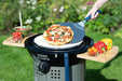 Cadac Pizza Lifter - BBQ Pizza Paddle lifestyle image of pizza cooked and being lifted off. The side of the Citi Chef 50 has red and yellow peppers and tomatoes one side and red tomatoes and a sauce in green bowl on the other wooded side table. Green gras