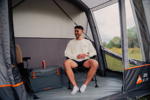 Vango Cove III Air Inflatable Drive Away Awning Smoke Grey - Low close up lifetsyle image of interior of awning with curtains open on right window side and man sat on chair with vango coolbox in the centre and can of drink on top. second chair with just a